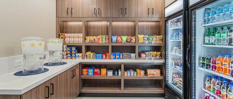 A kitchenette with a refrigerator stocked with beverages, a coffee machine, and shelves with snacks and condiments.