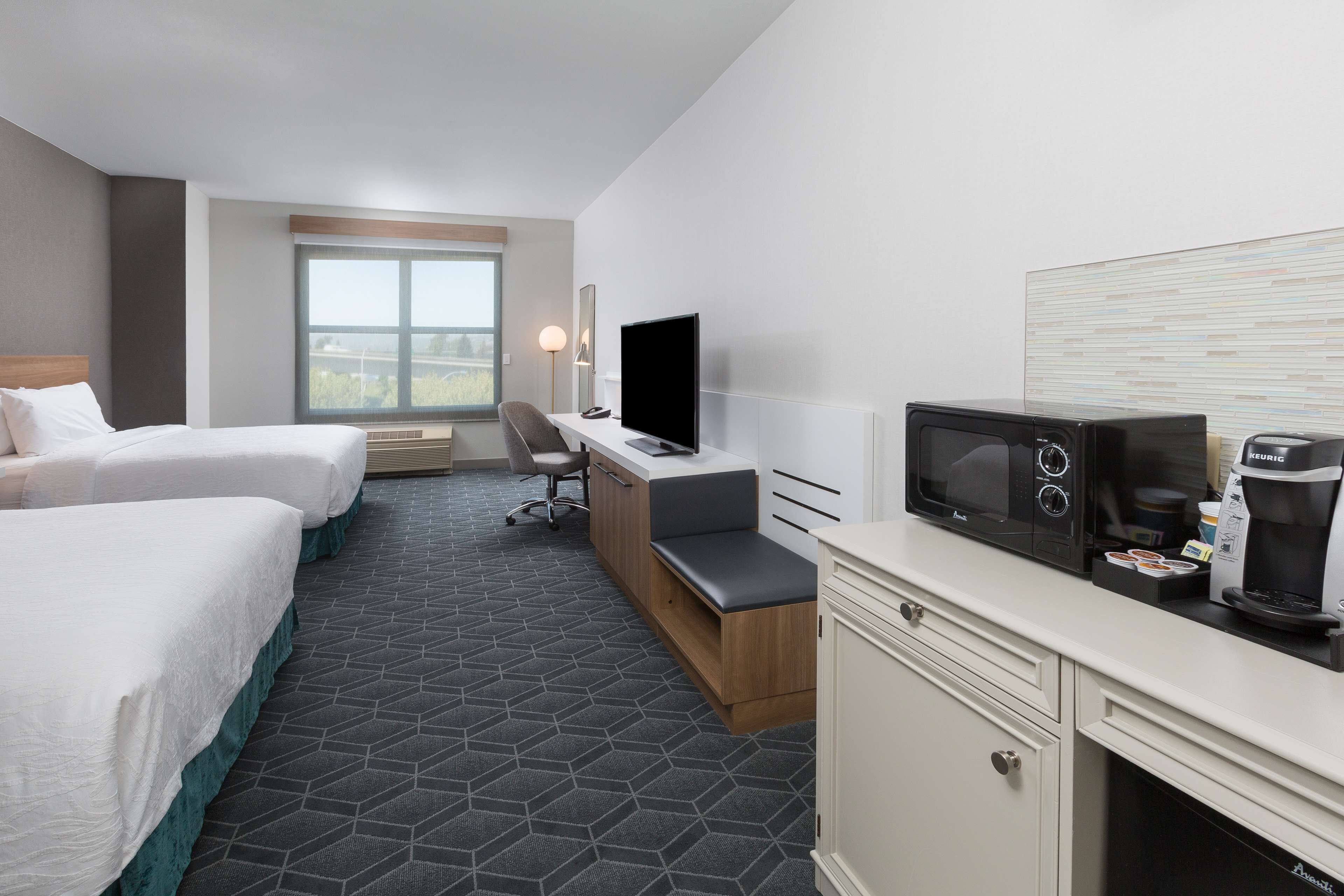 A hotel room with two beds, a desk with a flat-screen TV, a microwave, a coffee maker, and a large window offering a view.