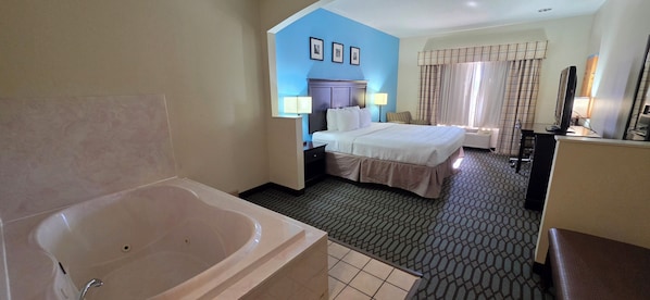 A hotel room with a large bathtub, a bed with two pillows, a desk with a computer, and a window with curtains.