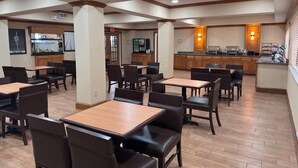 A hotel dining area with multiple tables and chairs, a kitchen area with buffet service, and a fish tank on the wall. - Wingate by Wyndham Lubbock TX South (Lubbock)