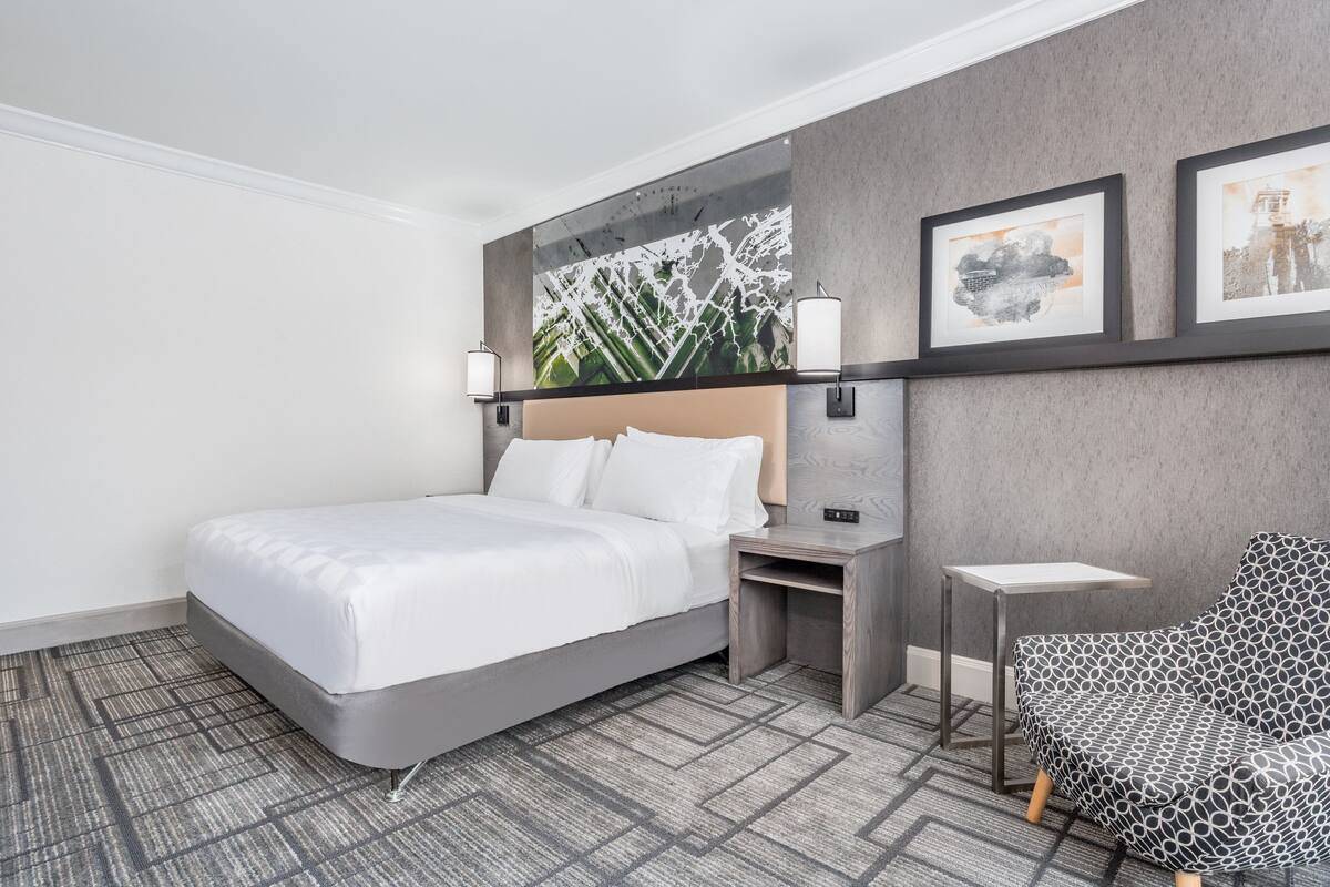 A modern hotel room with a large bed, a small desk, a chair, and artwork on the wall.