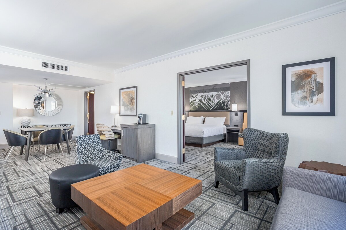 A hotel room with a wooden coffee table, patterned armchairs, a sofa, a bed with white linens, a desk with a lamp, and artwork on the walls.