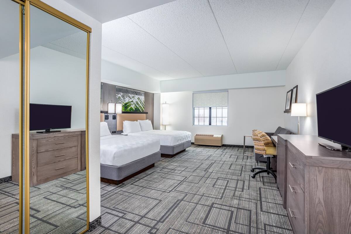 A hotel room with a large mirror, two beds, a desk, a TV, and a window with blinds.