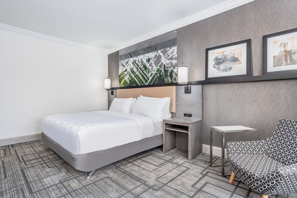 A modern hotel room with a large bed, a small desk, a chair, and artwork on the wall.