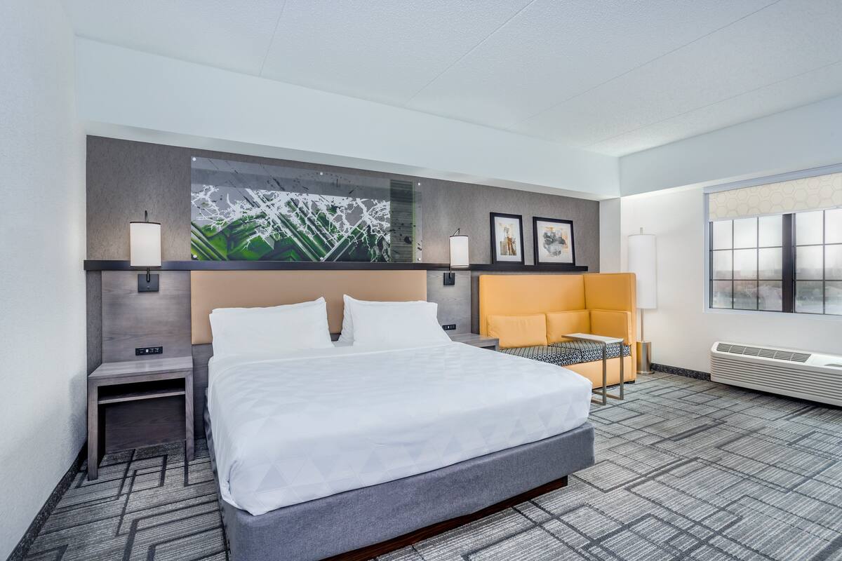A hotel room with a large bed, a desk, a chair, a wall-mounted TV, and artwork.
