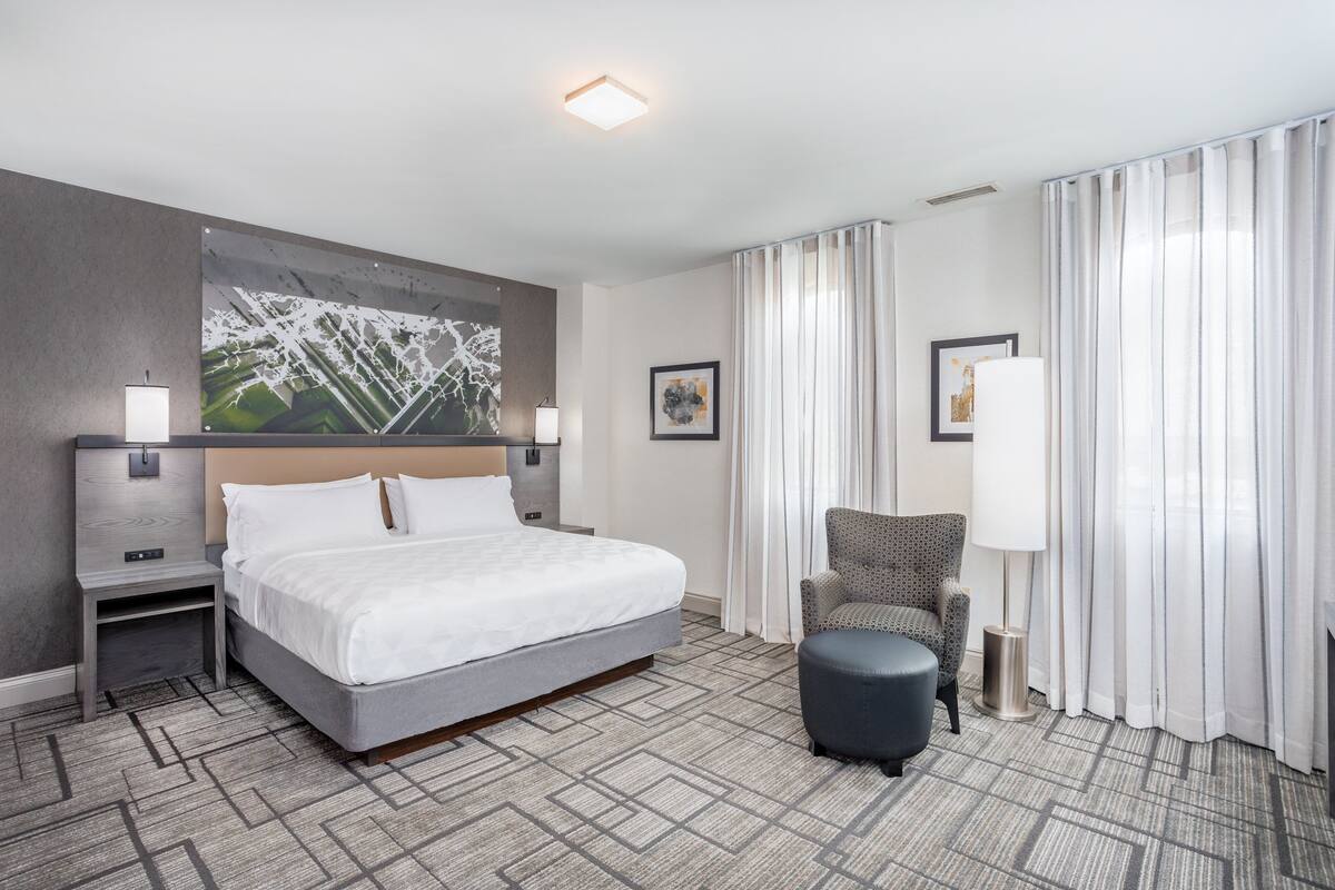A modern hotel room with a large bed, a chair, a small table, and a painting on the wall.
