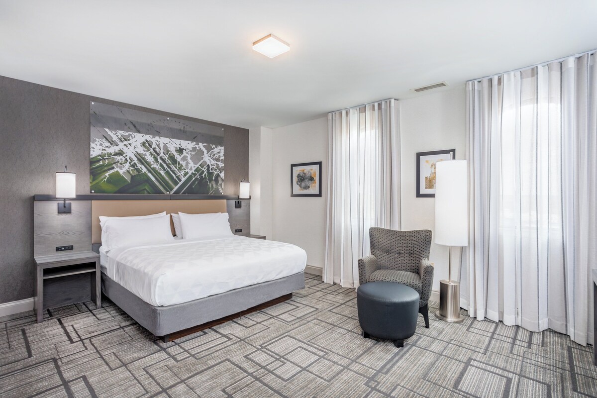 A hotel room with a large bed, a chair, a small table, and a painting on the wall.