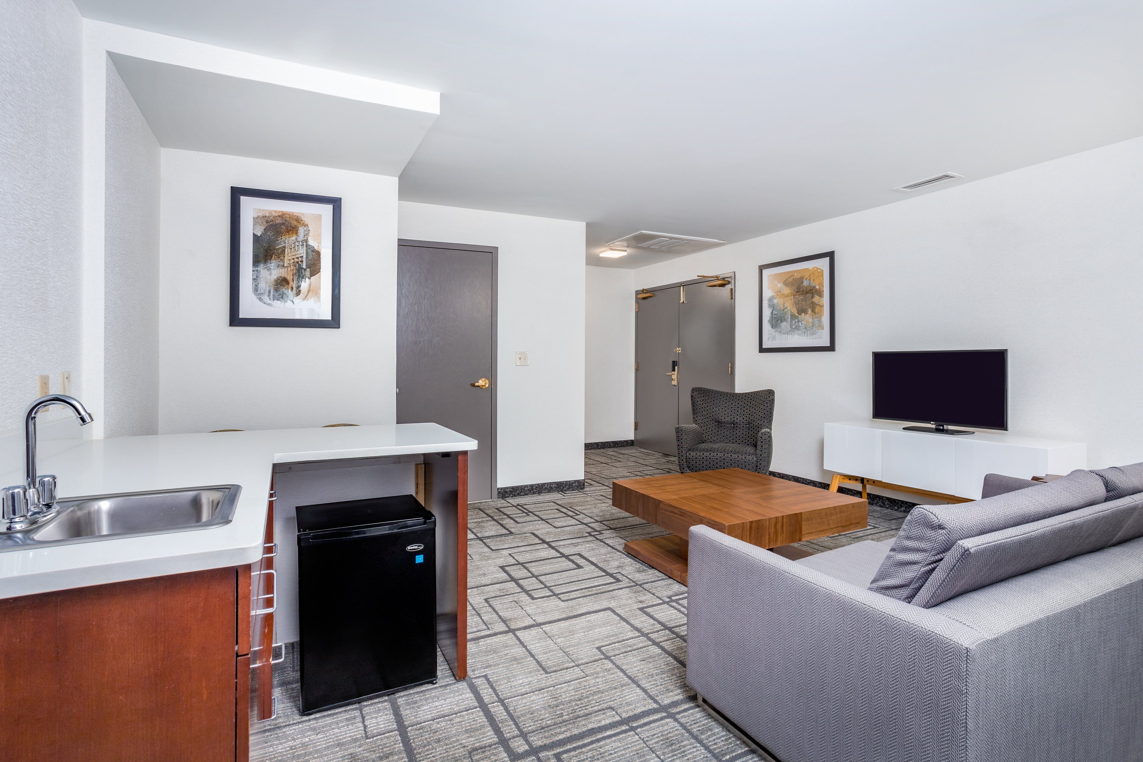 Una habitación de hotel moderna con kitchenette, sofá, televisión y obras de arte en las paredes.