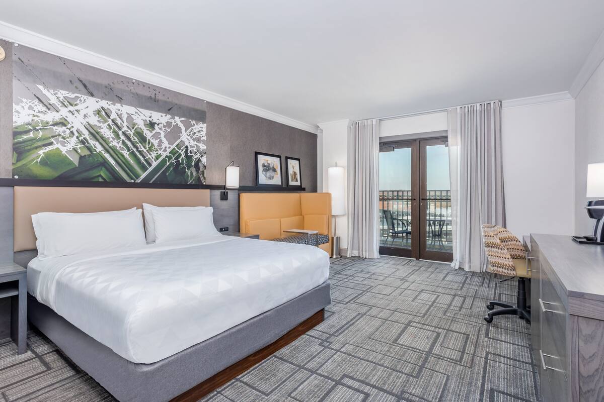 A hotel room with a large bed, a desk, a chair, a balcony, and a cityscape artwork on the wall.