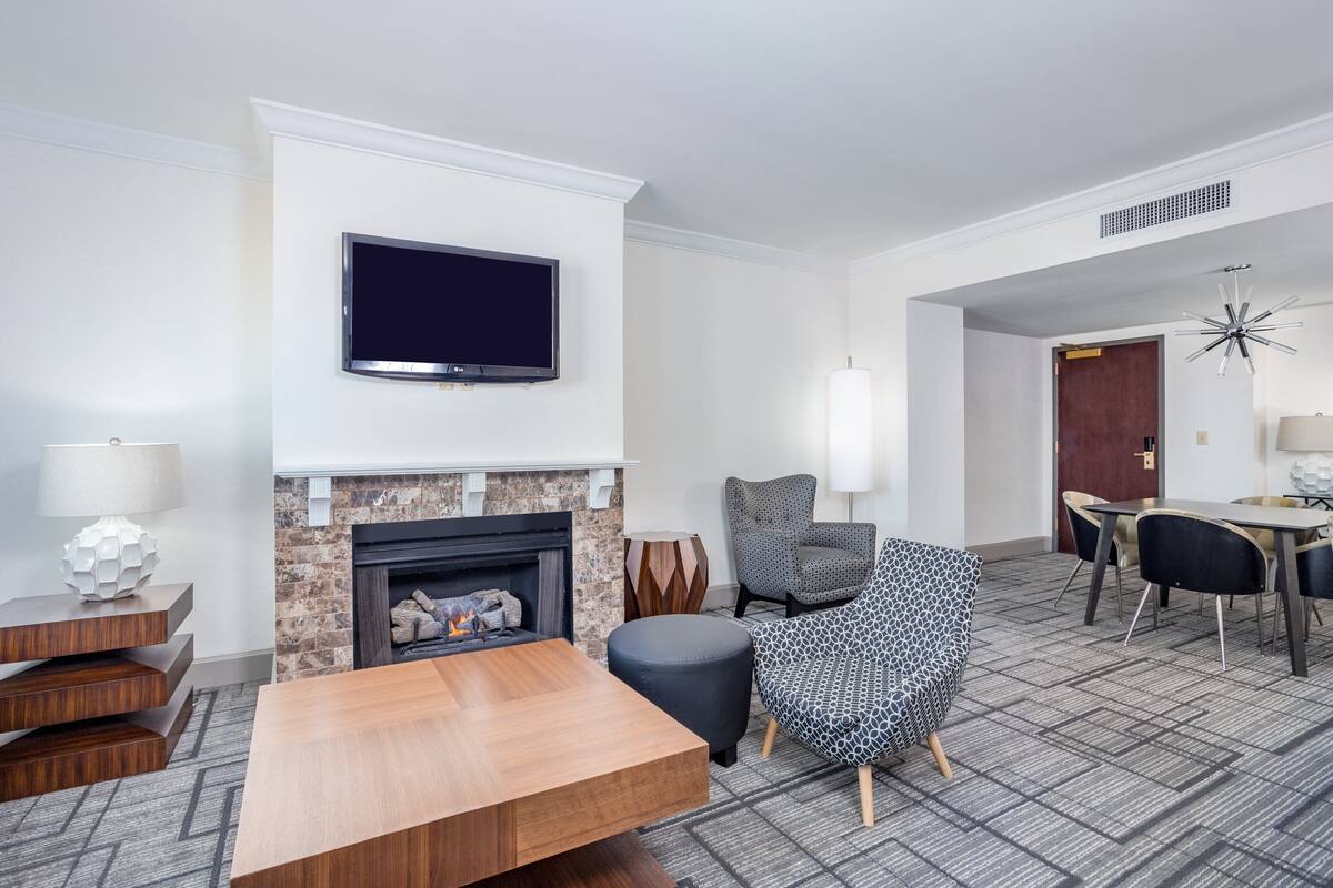 A modern hotel room with a fireplace, a flat-screen TV mounted above it, a dining area with a table and chairs, and a patterned armchair.