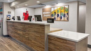 Balcão de recepção de hotel com design em painel de madeira, uma estação de check-in digital e um letreiro com a mensagem 'Welcome to Hampton'.