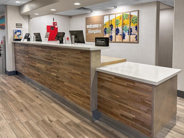 A hotel reception desk with a wooden panel design, a digital check-in station, and a 'Welcome to Hampton' sign.