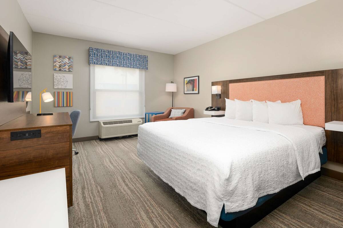 A hotel room with a large bed, a desk with a lamp, a chair, a window with blinds, and a wall-mounted TV.