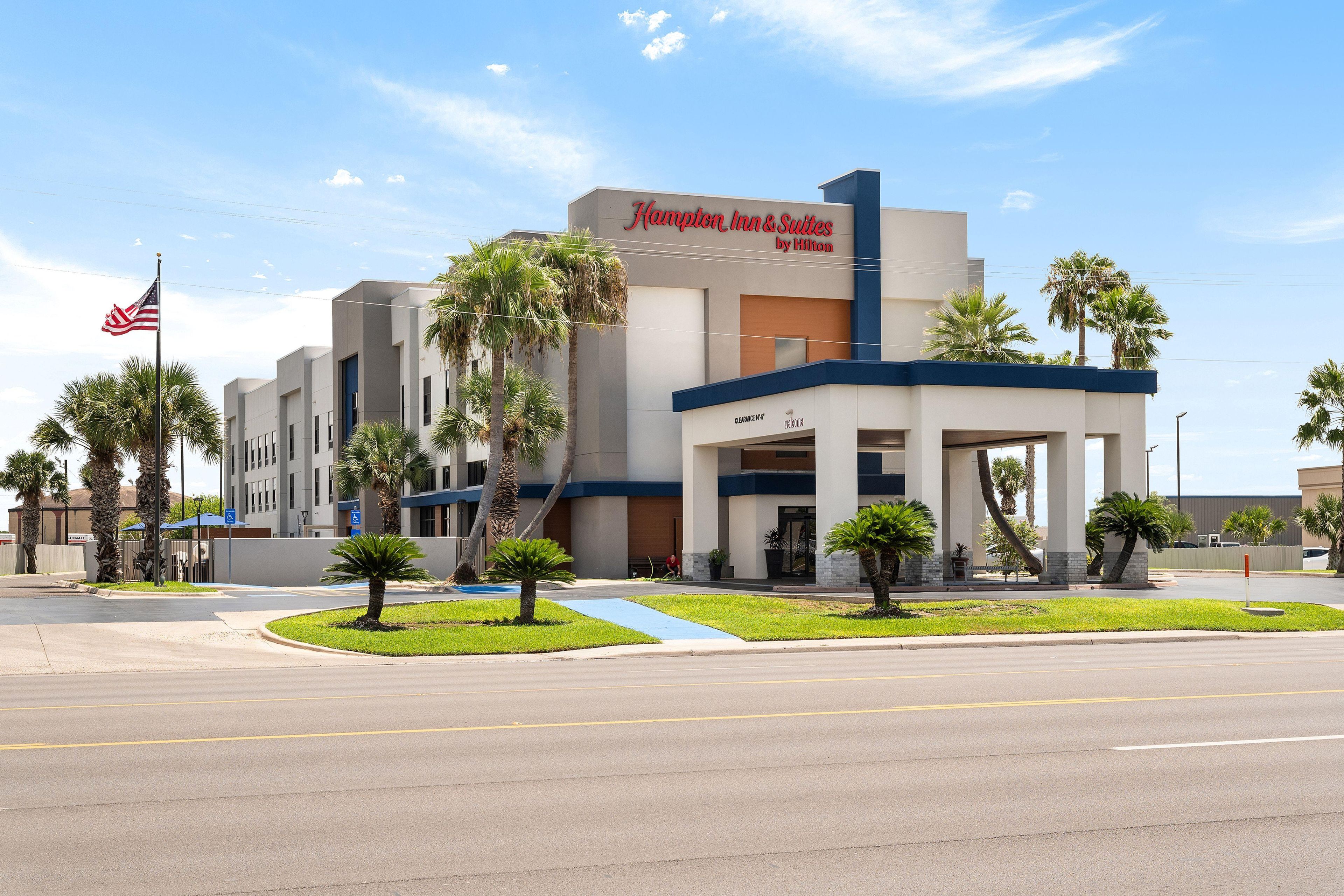 Un hôtel Hampton Inn & Suites de la chaîne Hilton, doté d’une façade moderne, de palmiers et surplombant un ciel dégagé.
