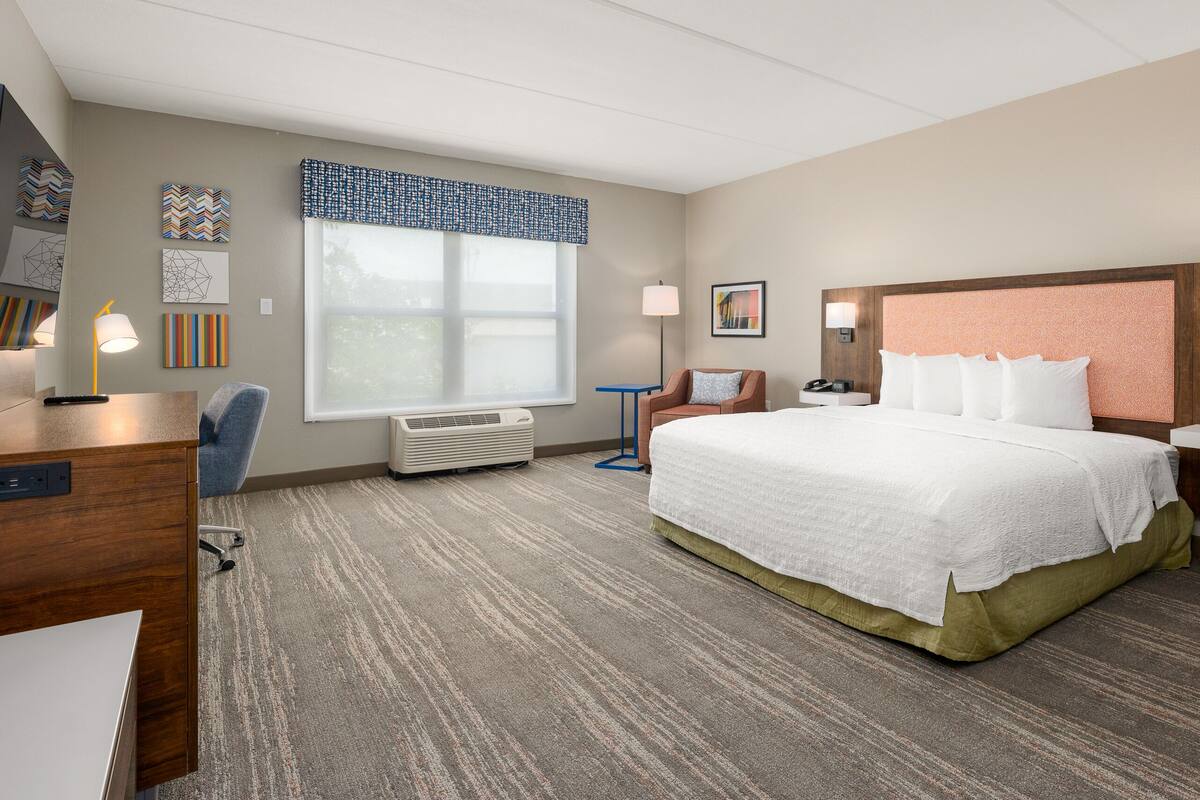 A hotel room with a large bed, a desk, a chair, a window with blinds, a wall-mounted heater, and a nightstand with a lamp.