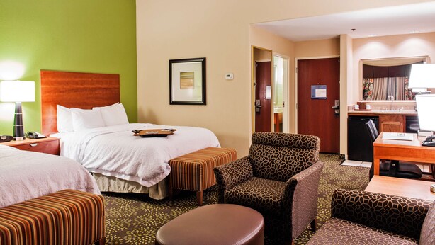 A hotel room with a bed, two ottomans, a chair, a desk, and a kitchenette. - Hampton Inn & Suites Harlingen (Harlingen)