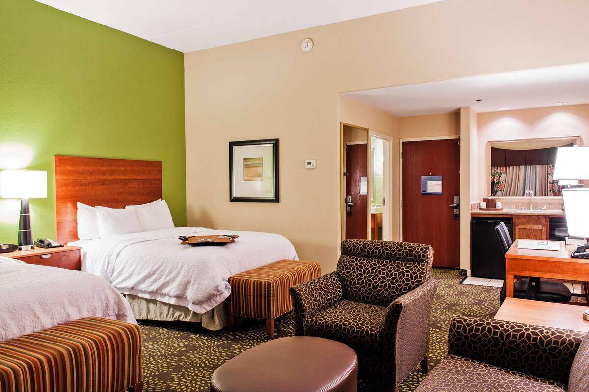 A hotel room with a bed, two ottomans, a chair, a desk, and a kitchenette.