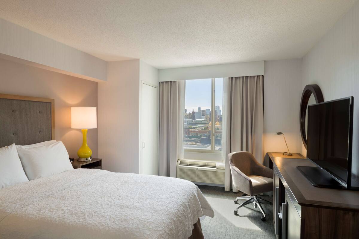 A hotel room with a bed, a desk with a TV, a chair, a lamp, and a large window with a city view.