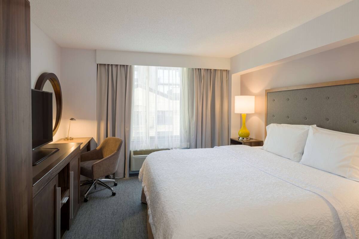 A hotel room with a large bed, a desk with a chair, a television, a lamp, and a window with curtains.