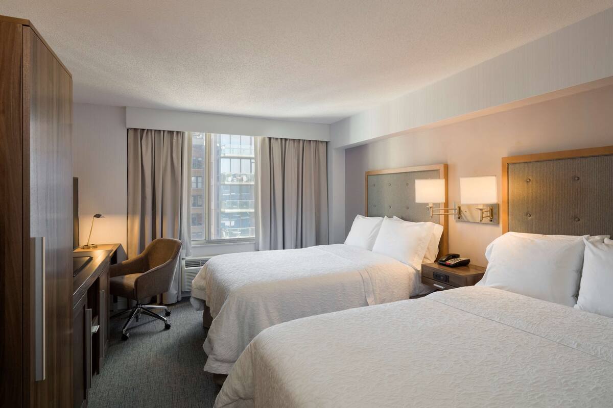 A hotel room with two beds, a desk, a chair, a window with curtains, and a nightstand with a lamp.