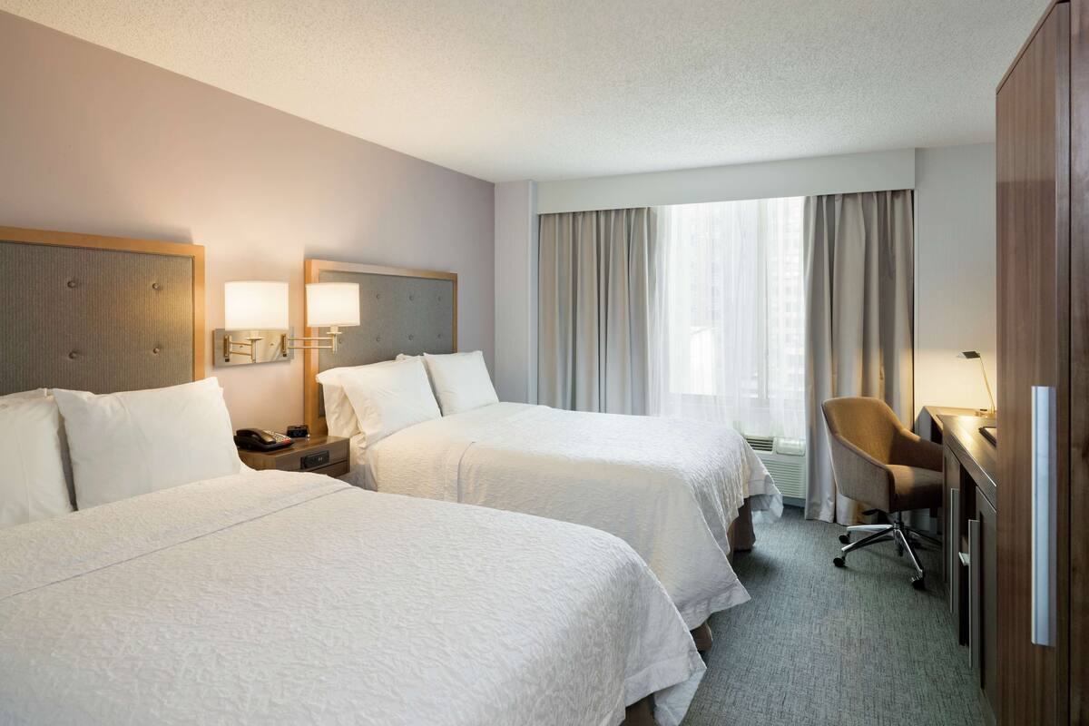 A hotel room with two beds, a desk, a chair, and a large window with curtains.