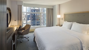 A hotel room with a large bed, a desk with a chair, a window with curtains, and a view of the cityscape. - Hampton Inn Manhattan-Chelsea (New York)