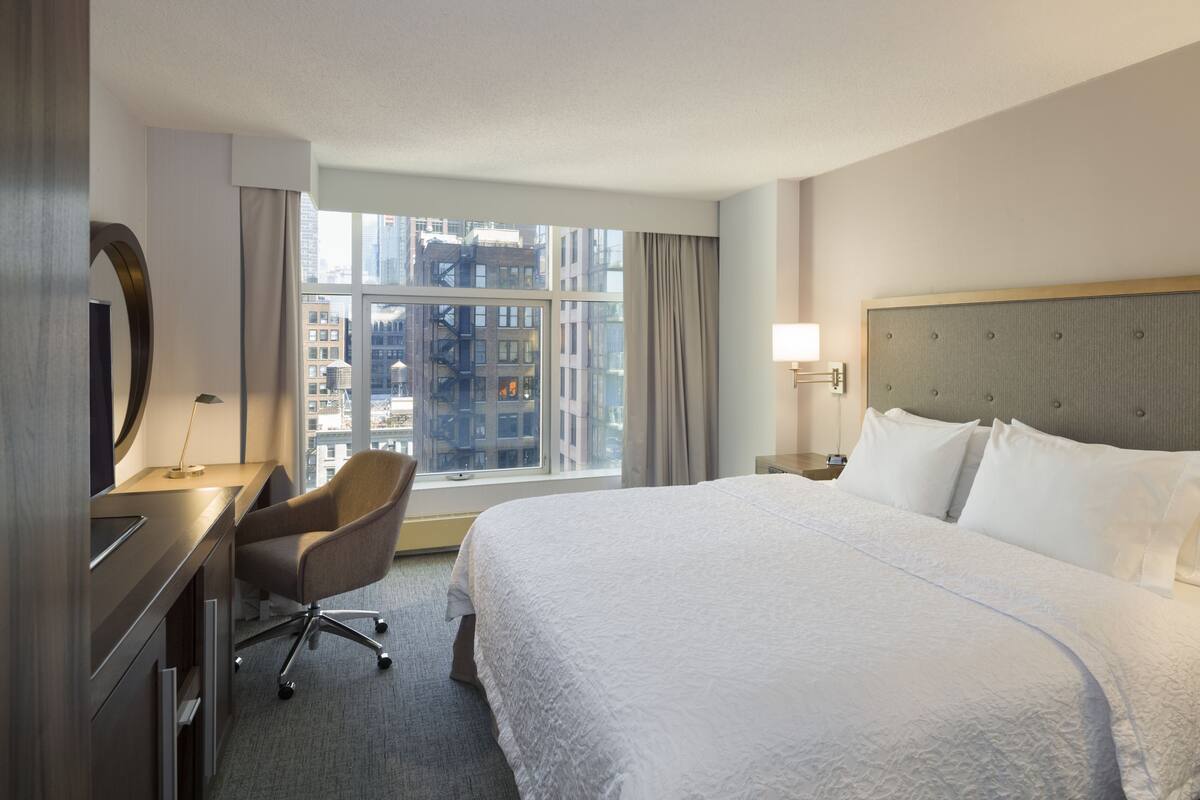 A hotel room with a large bed, a desk with a chair, a window with curtains, and a view of the cityscape.