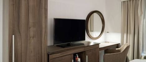 A hotel room with a wooden wardrobe, a flat-screen TV, a desk with a lamp, a chair, and a bed.