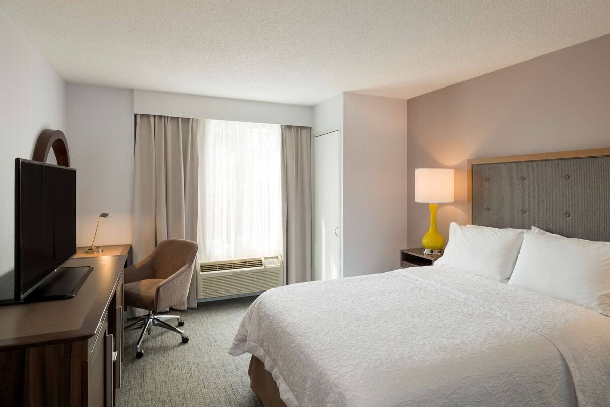 A hotel room with a bed, a desk with a computer, a chair, a TV, a lamp, and a window with curtains.