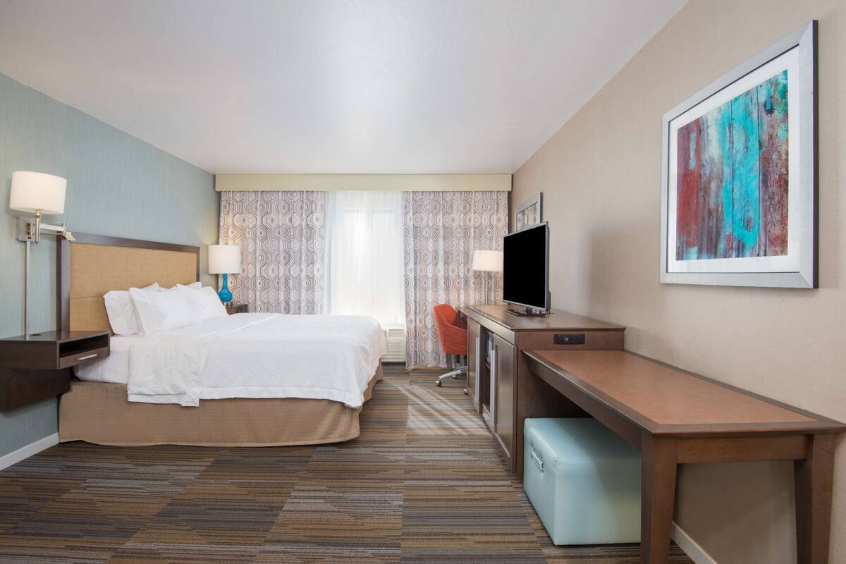 A hotel room with a bed, desk, TV, and a painting on the wall.