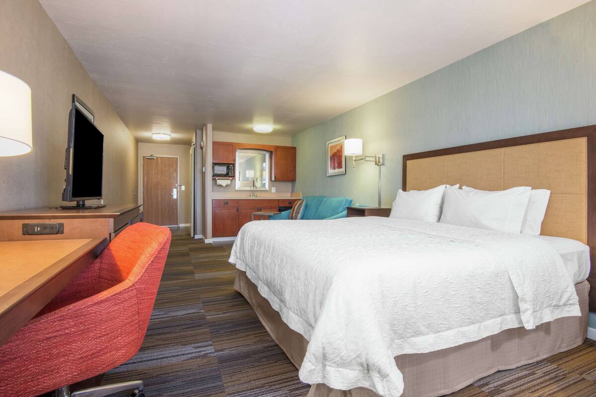 A hotel room with a bed, desk, chair, and kitchenette.