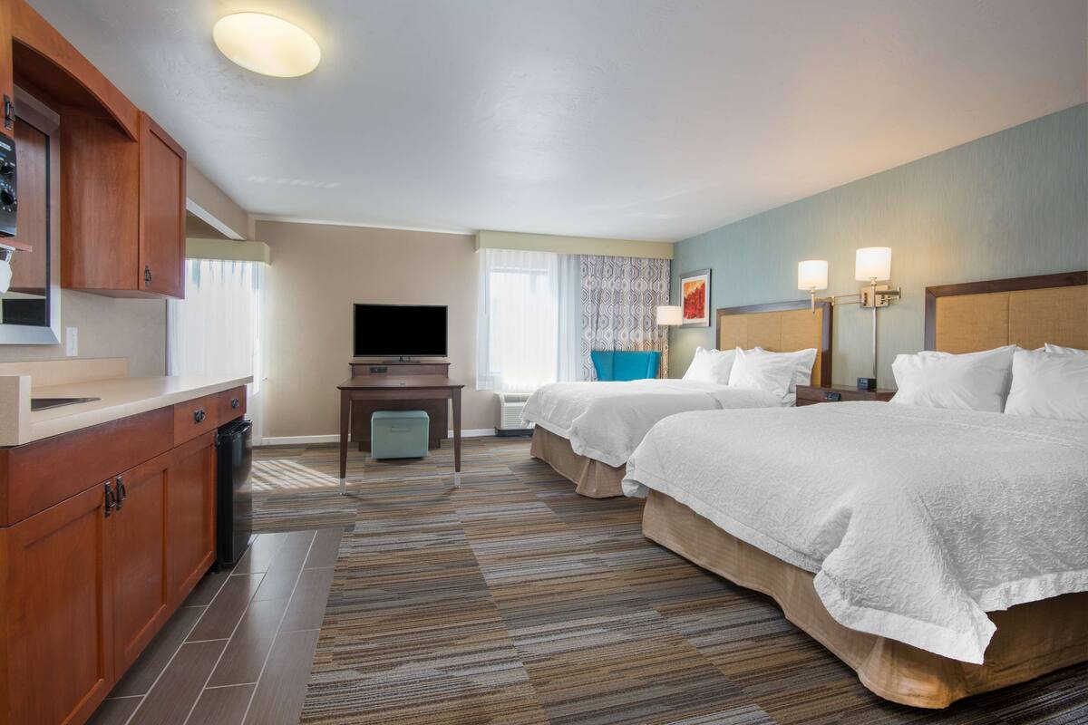 A hotel room with two beds, a TV, a desk, and a kitchenette.
