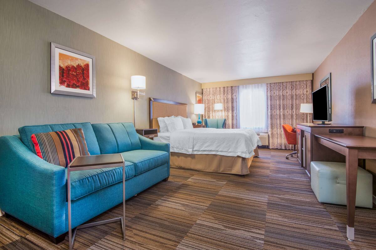 A hotel room with a blue sofa, a bed with white linens, a desk with a television, and a patterned curtain.