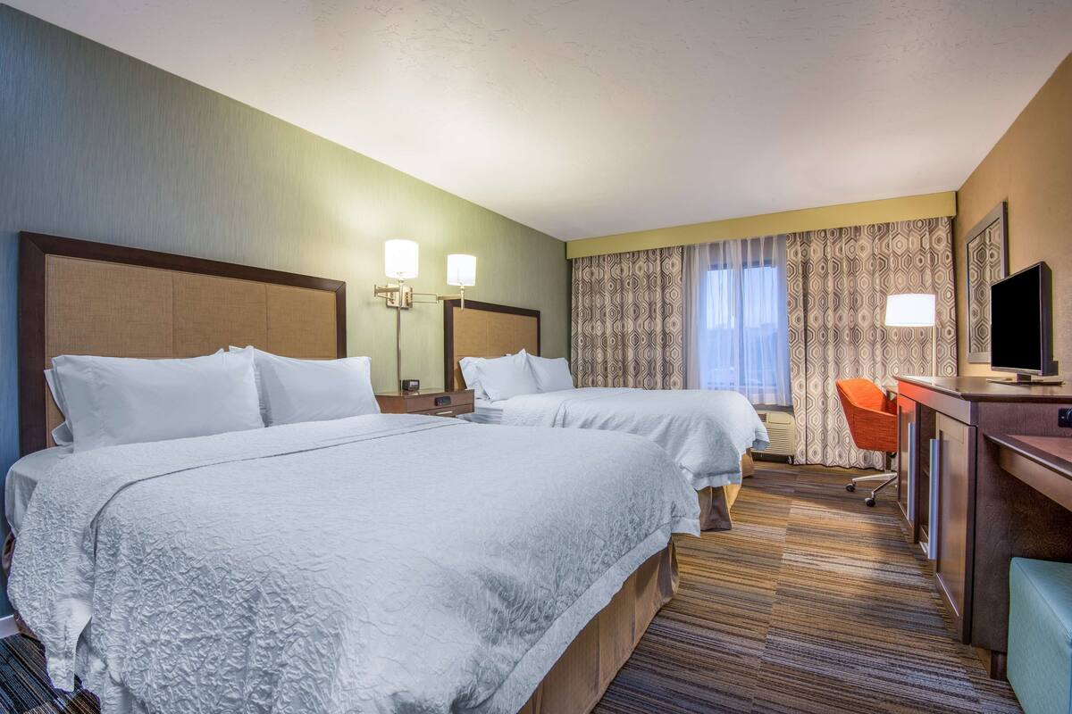A hotel room with two beds, a desk with a TV, a chair, and a large window with curtains.