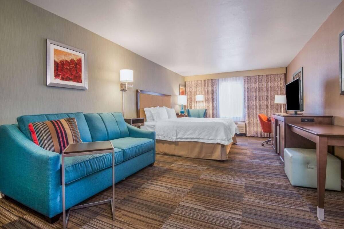 A hotel room with a bed, a sofa, a desk, a TV, and a painting on the wall.