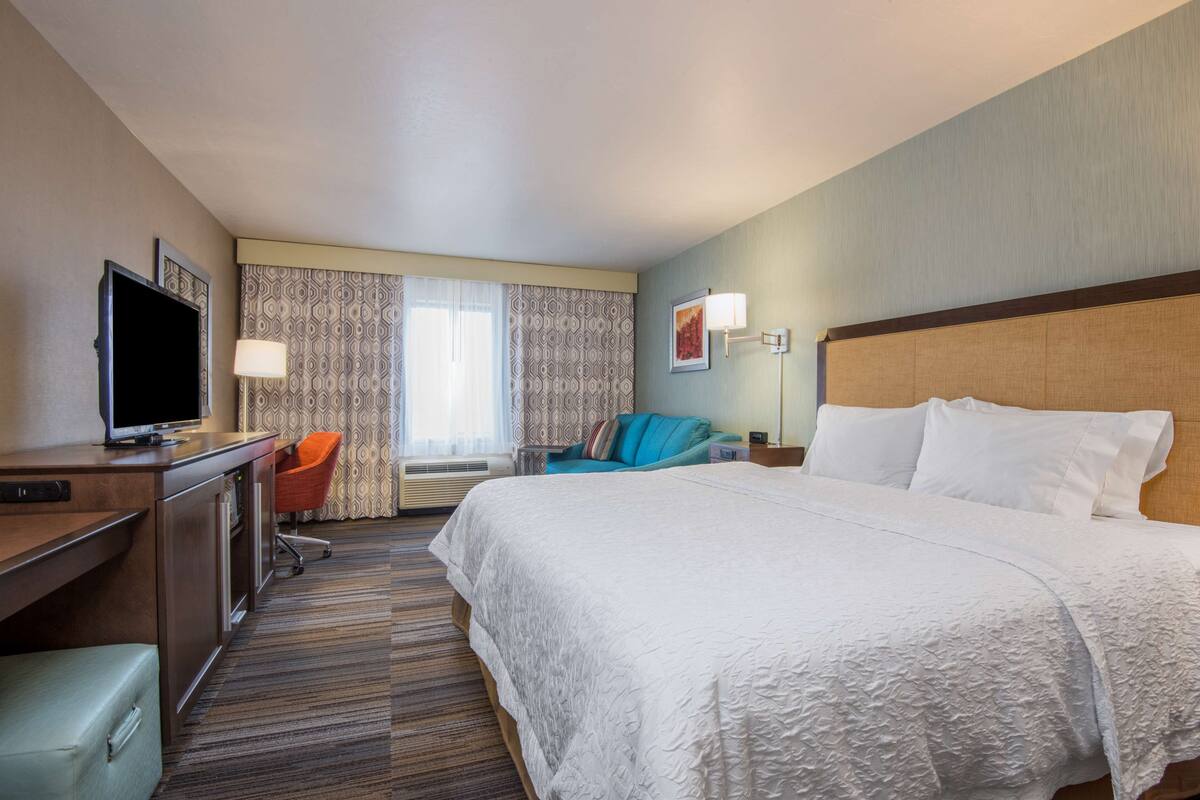 A hotel room with a large bed, a desk with a flat-screen TV, a chair, and a window with curtains.
