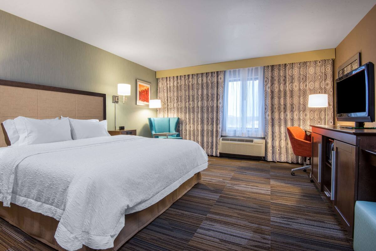 A hotel room with a large bed, a desk with a television, a chair, and a window with curtains.