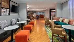 Een hotellobby met een zithoek met banken, poefs en tafels. Achterin is een receptiebalie en een bar zichtbaar.