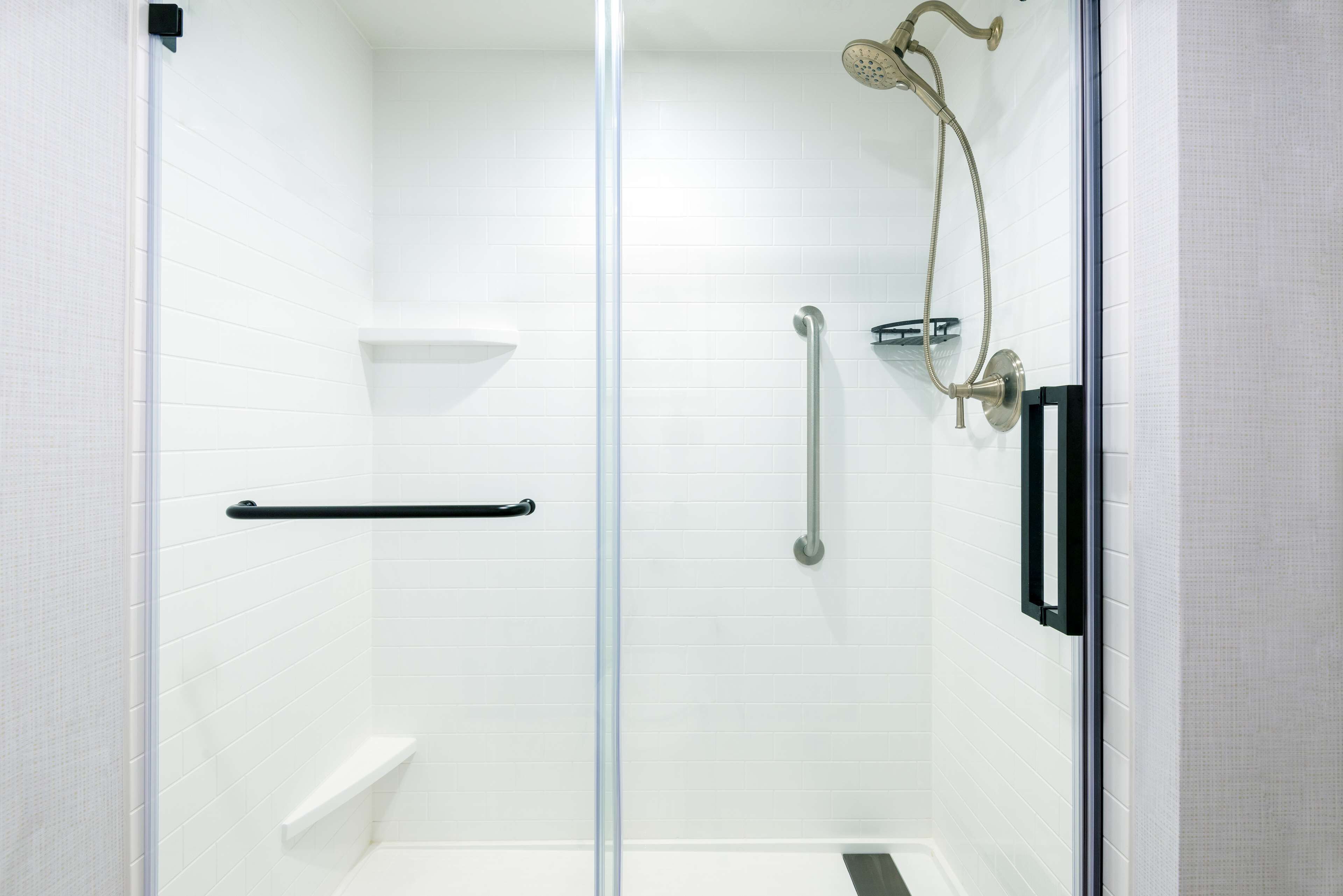 A glass-enclosed shower with white tiled walls, a handheld showerhead, and a handheld shower control.