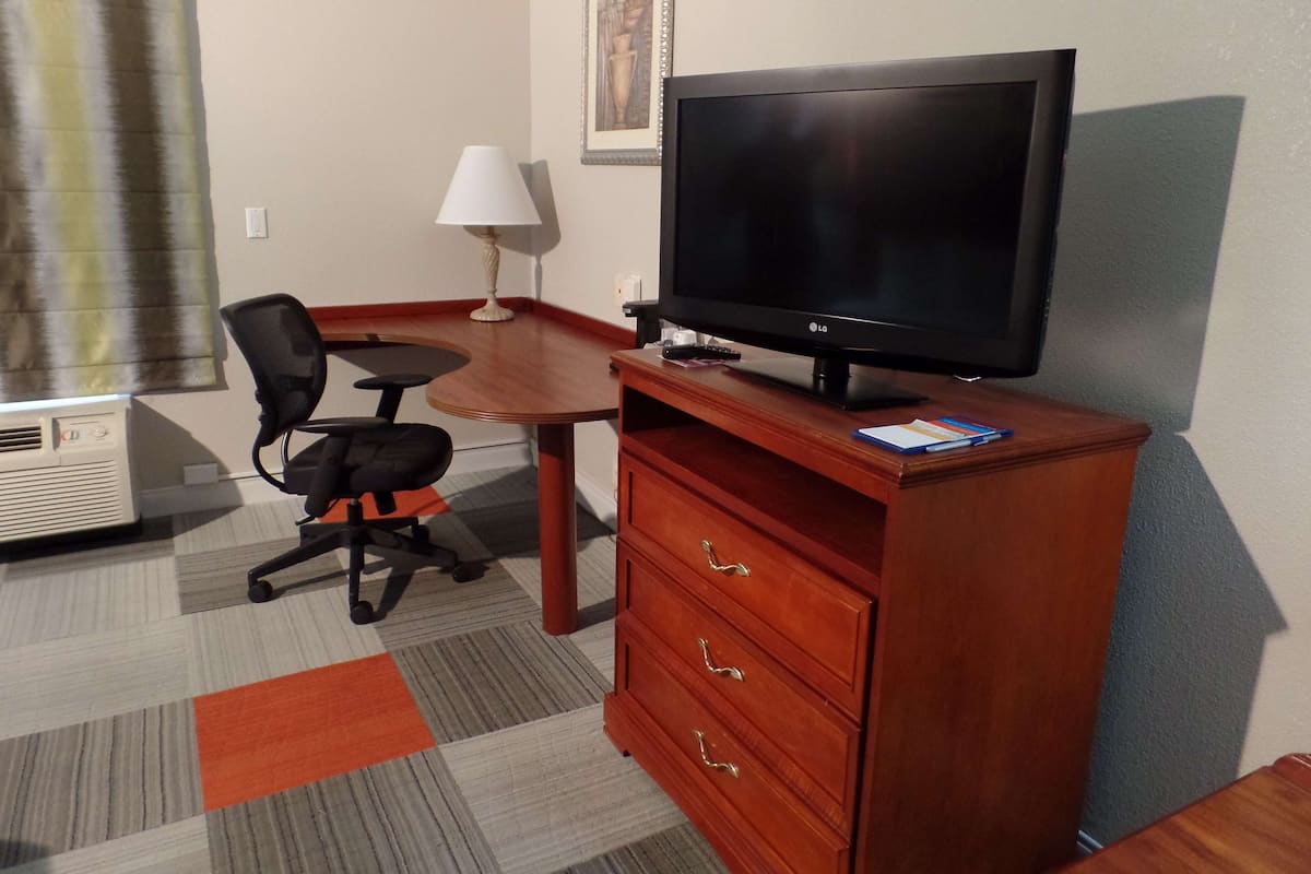 Quarto de hotel com escrivaninha de madeira, cadeira de escritório preta, televisão sobre suporte de madeira, luminária e um pequeno aparelho de ar condicionado.