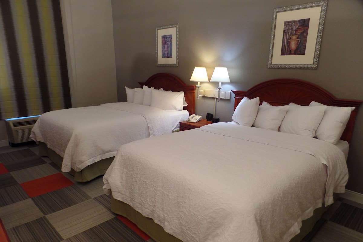 Quarto de hotel com duas camas, uma mesa de cabeceira com abajur e um quadro na parede.