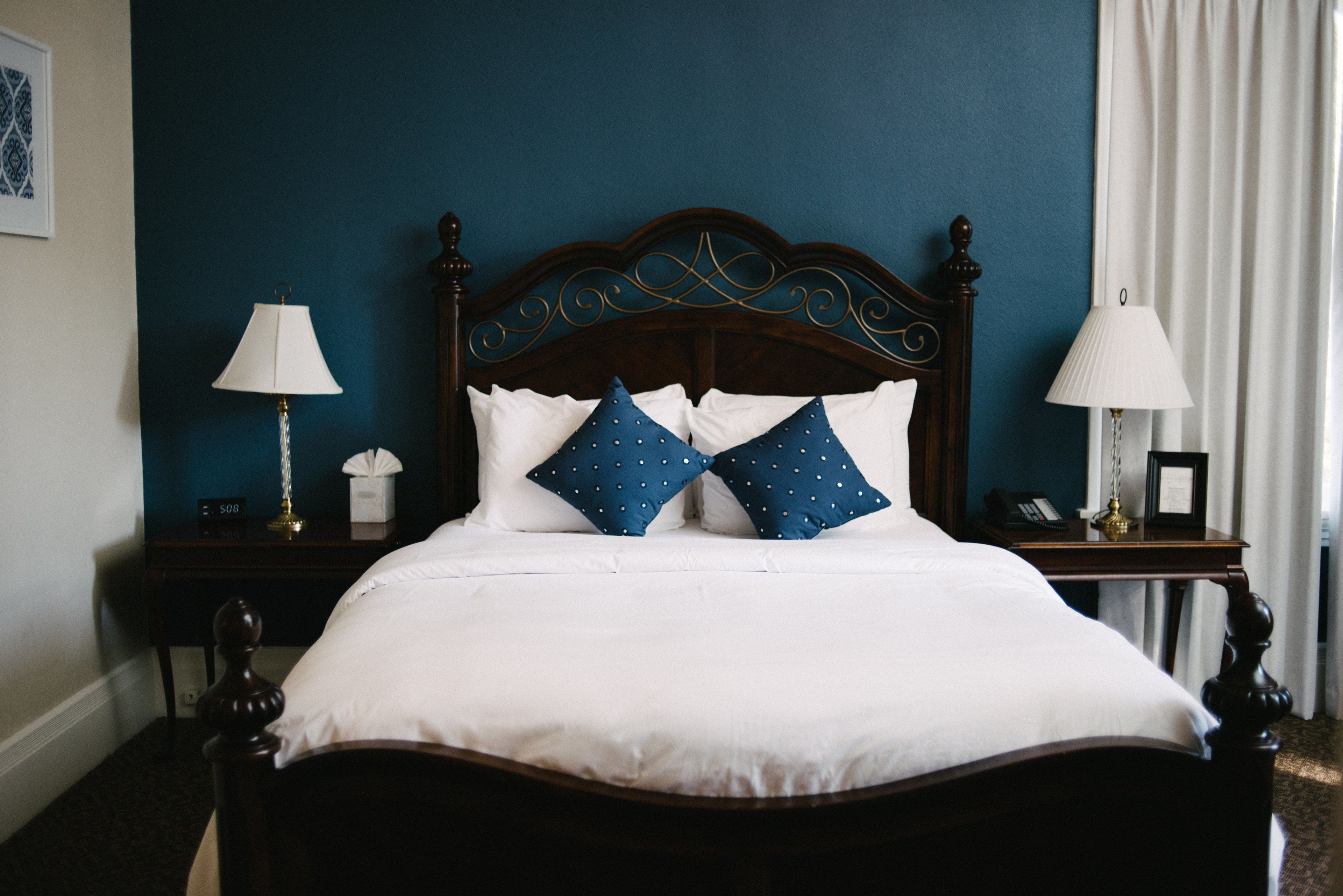 Un lit bien fait, avec du linge de lit blanc et des oreillers bleus, une tête de lit en bois et deux tables de chevet ornées de lampes et d’une horloge.