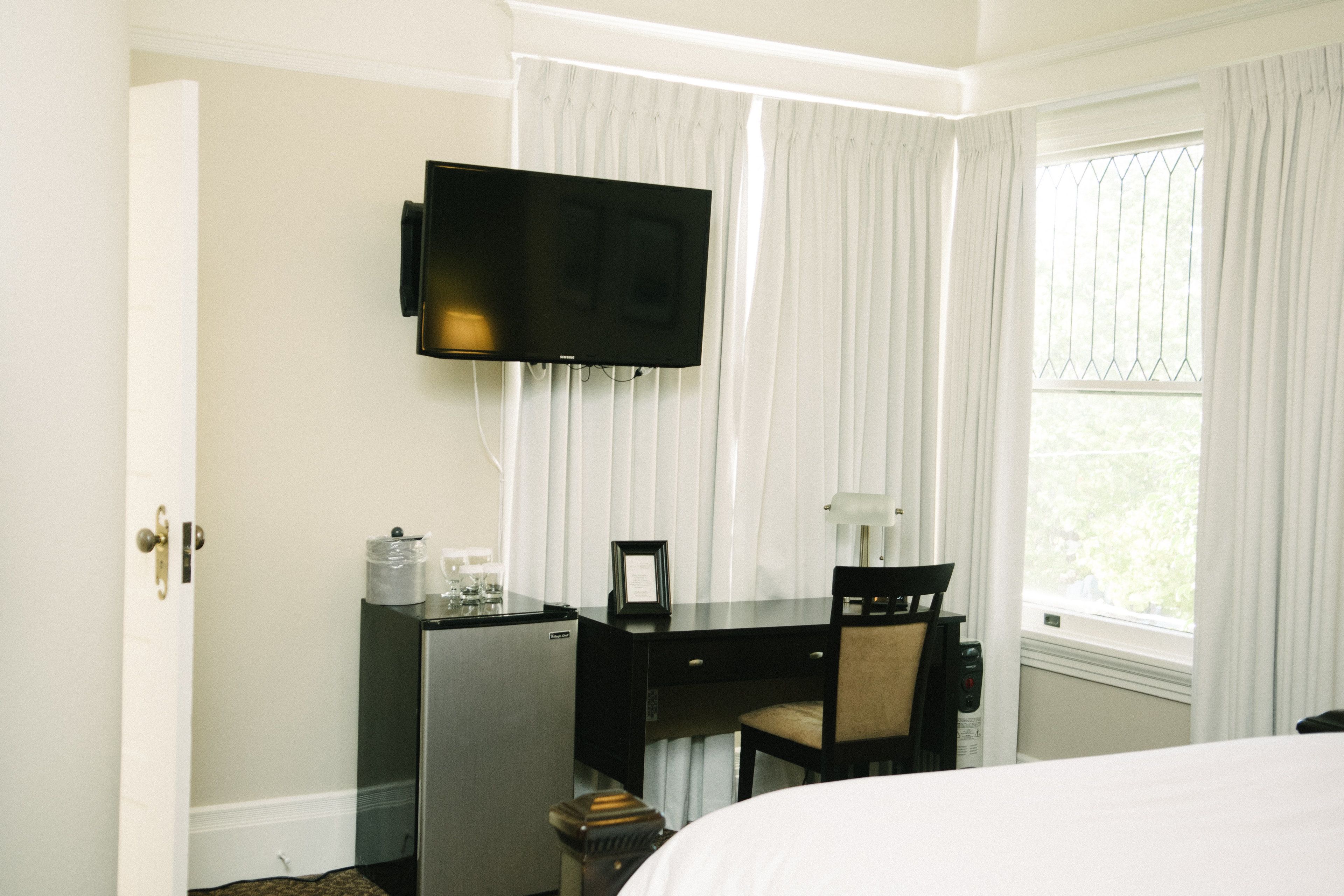 Une chambre d’hôtel comprenant un lit, un bureau avec une chaise, un téléviseur fixé au mur, une fenêtre avec des rideaux et une porte.