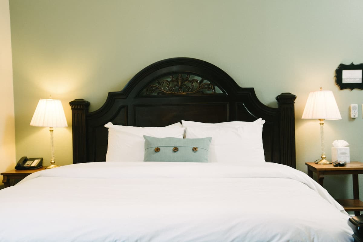 A neatly made bed with white linens, two bedside lamps, a telephone, and a small table with a lamp and tissues.