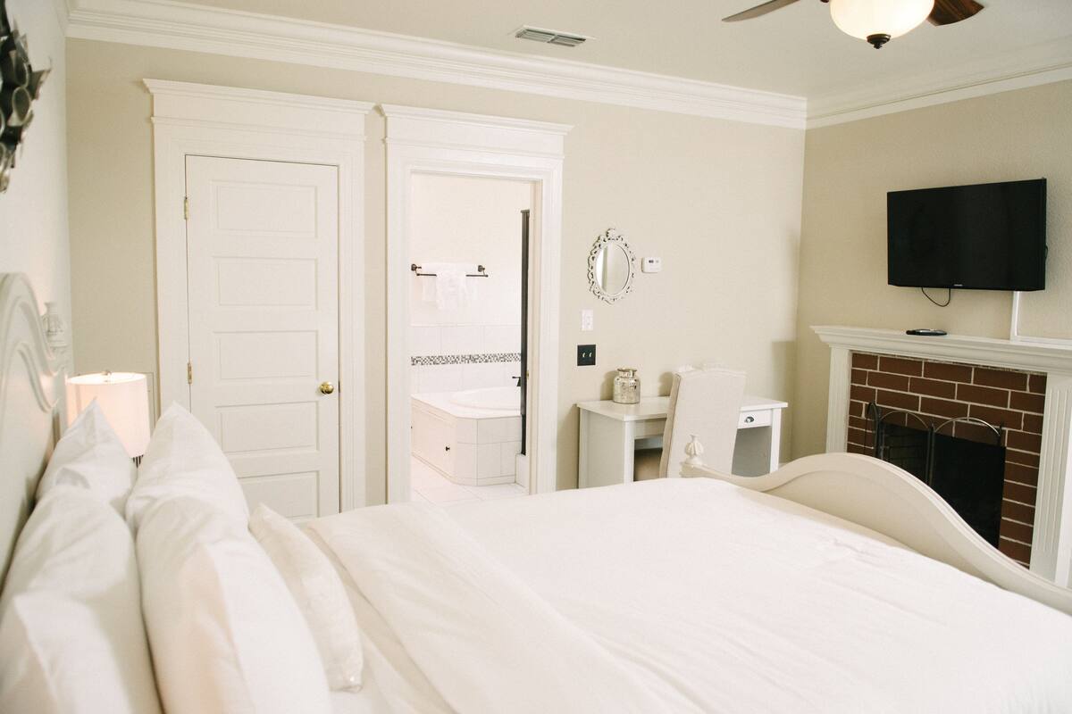 A bedroom with a bed, a television mounted above a fireplace, and a bathroom visible through an open door.