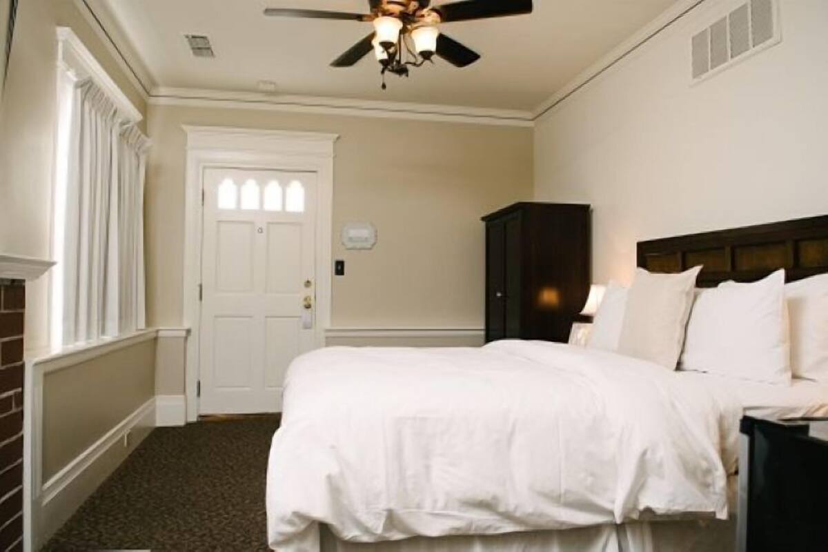 A hotel room with a bed, a ceiling fan, a dark wooden cabinet, and a white door.
