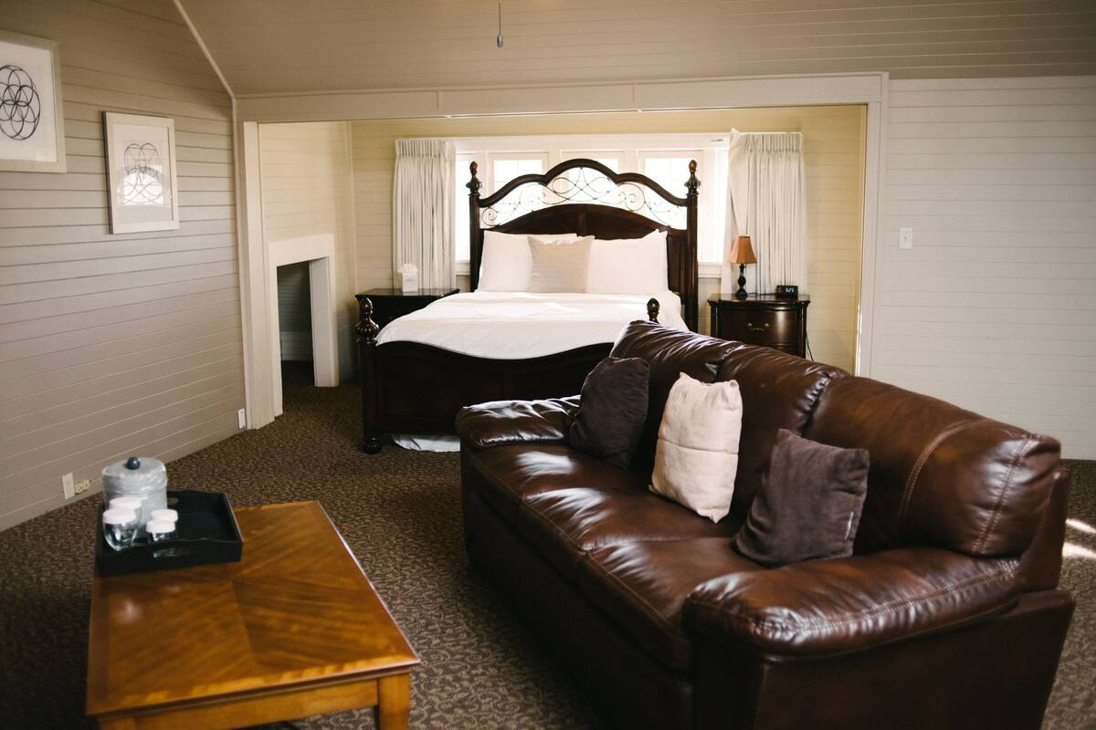 A bedroom with a large bed, a leather sofa, a wooden side table, and a fireplace.