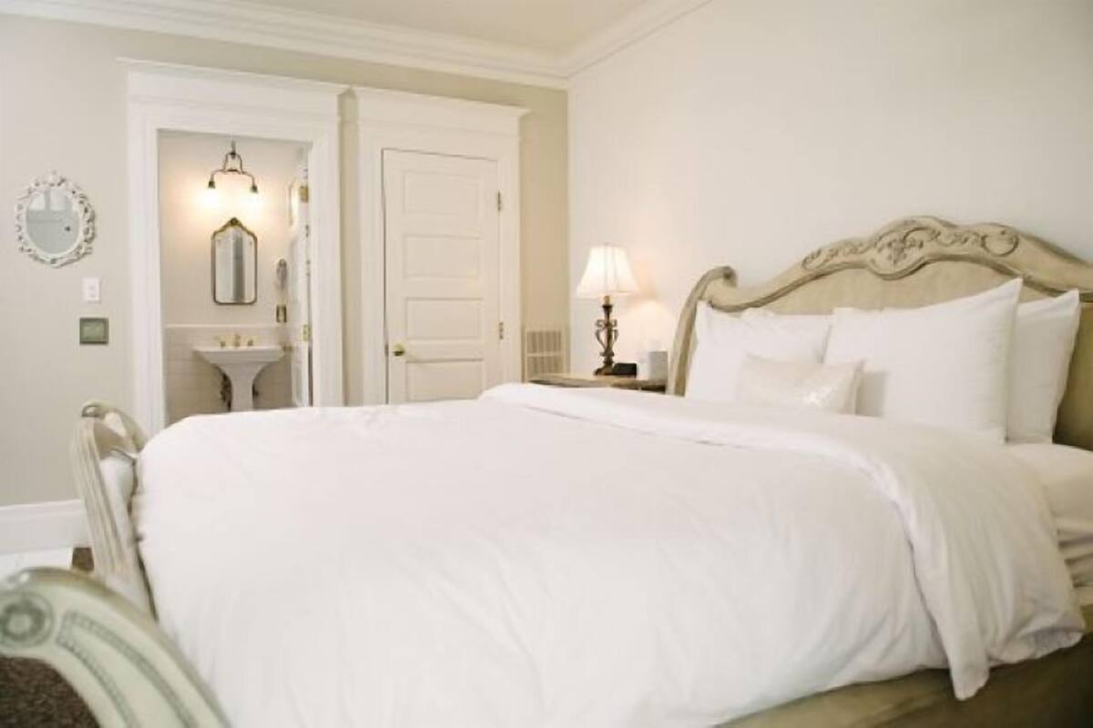 A neatly made bed with white linens, a bedside table with a lamp, and a door leading to a bathroom with a mirror and sink.