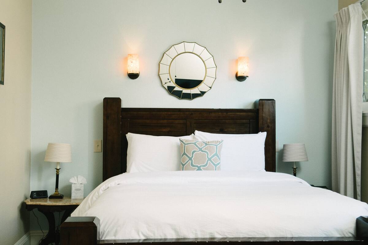 A neatly made bed with white linens, a wooden headboard, two bedside lamps, a wall-mounted mirror, and a bedside table with a lamp and tissues.