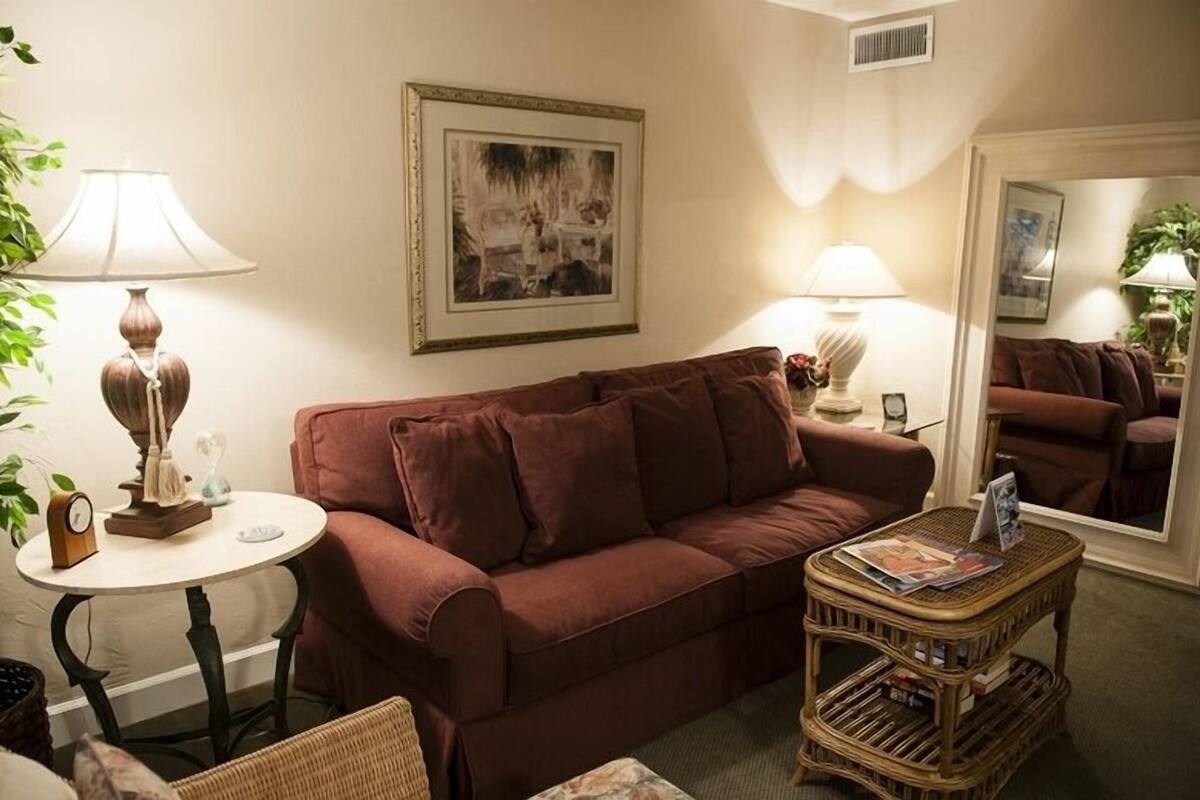 A living room with a brown sofa, two lamps, a round table with a clock, a small wicker side table with magazines, and a framed picture on the wall.